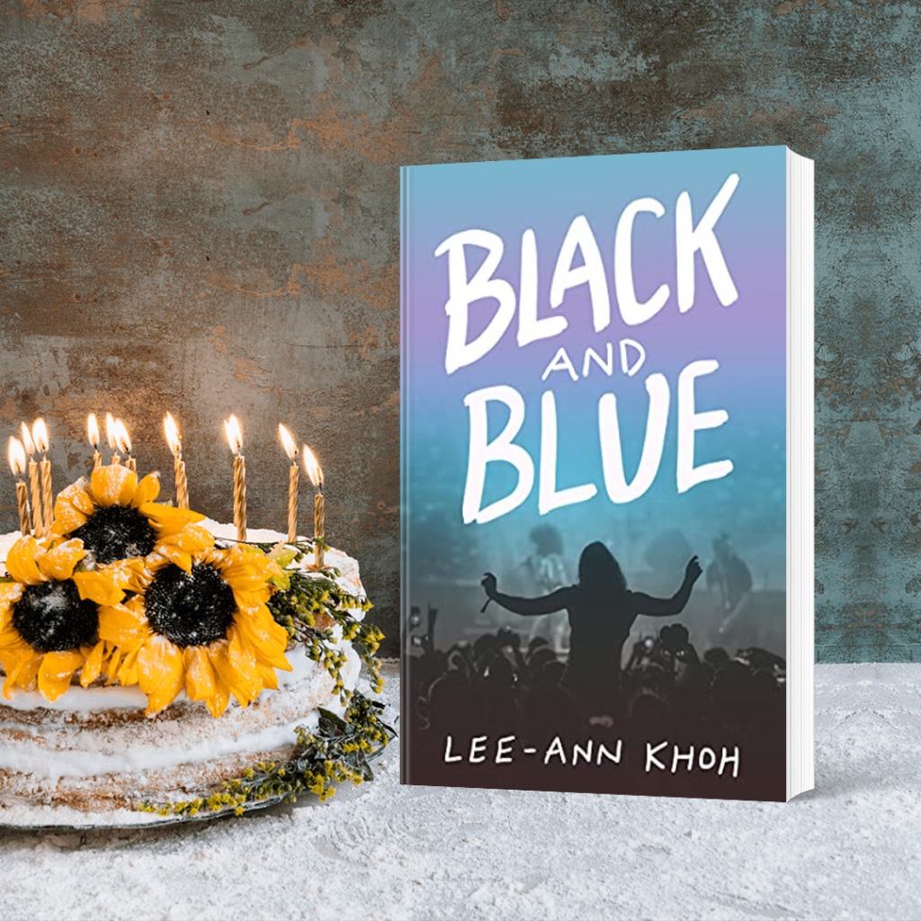 A paperback copy of Black and Blue by Lee-Ann Khoh stands next to a powdered sugar layer cake decorated with flowers and birthday candles.